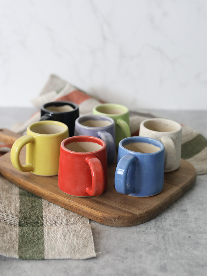Eight handmade ceramic espresso mugs arranged on a wooden board, viewed from a slight overhead angle showing the full colour range of yellow, black, red, purple, spring green, blue, and speckled white, each containing espresso, styled with linen cloths against a marble background