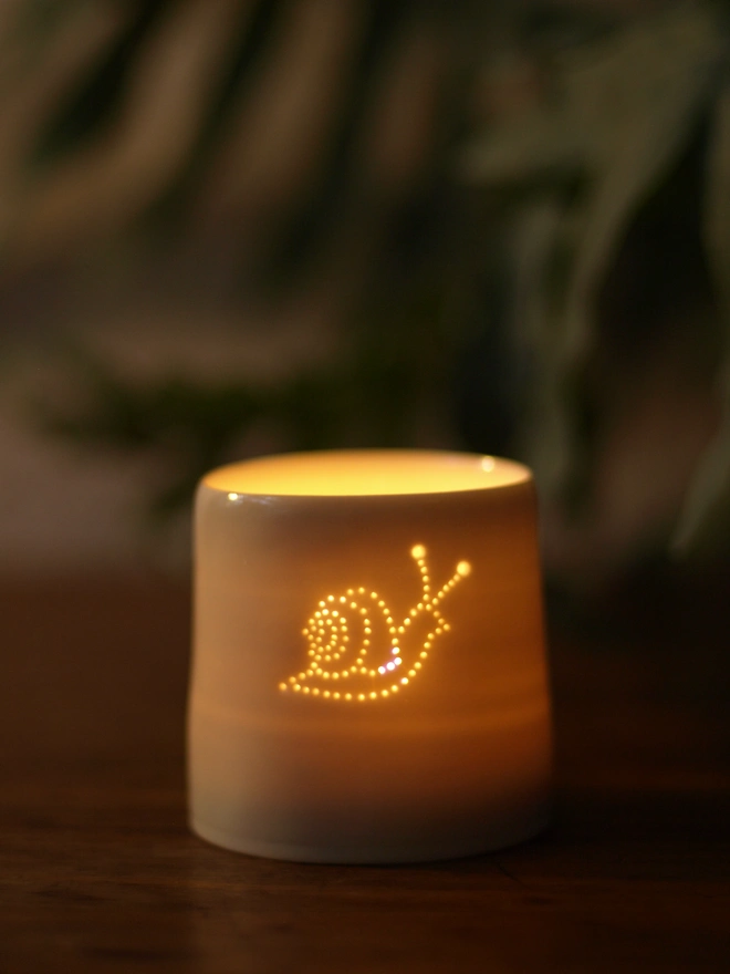 Single porcelain tealight holder, with small holes pierced in the clay, allowing the light to shine through the hole and form an image. The dots are in the pattern of a snail facing right, showing the curly shell.