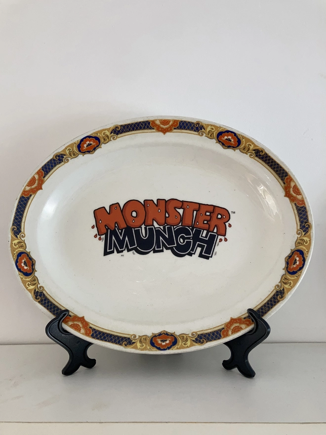 oval shaped vintage plate with the 'Monster Munch' logo in the centre. The rim of the plate is orange and dark blue, and the logo has been created to reflect these colours.