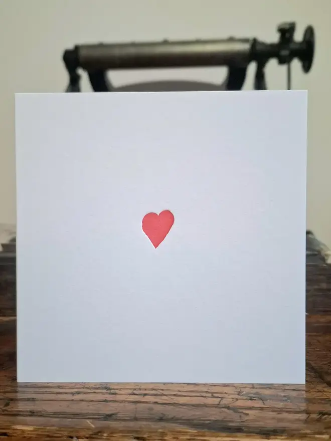 Small Red Heart Letterpress Handprinted Greetings Card, a white card with a small red heart sitting above a wooden surface. 
