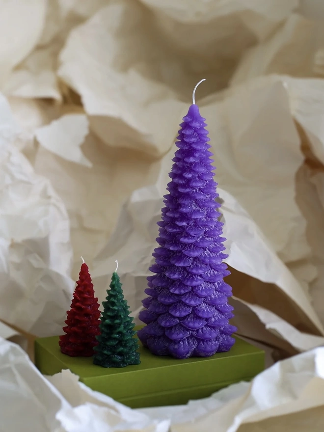 Large Christmas Tree Candle. a large purple christmas tree shaped candle stands on a green box, nestled among crumpled cream tissue paper. a smaller red and a smaller dark green tree candle sit in front of it for scale, both with visible wicks. the textured, layered pine branch design and slightly variegated wax give all three candles a realistic, hand-poured finish.