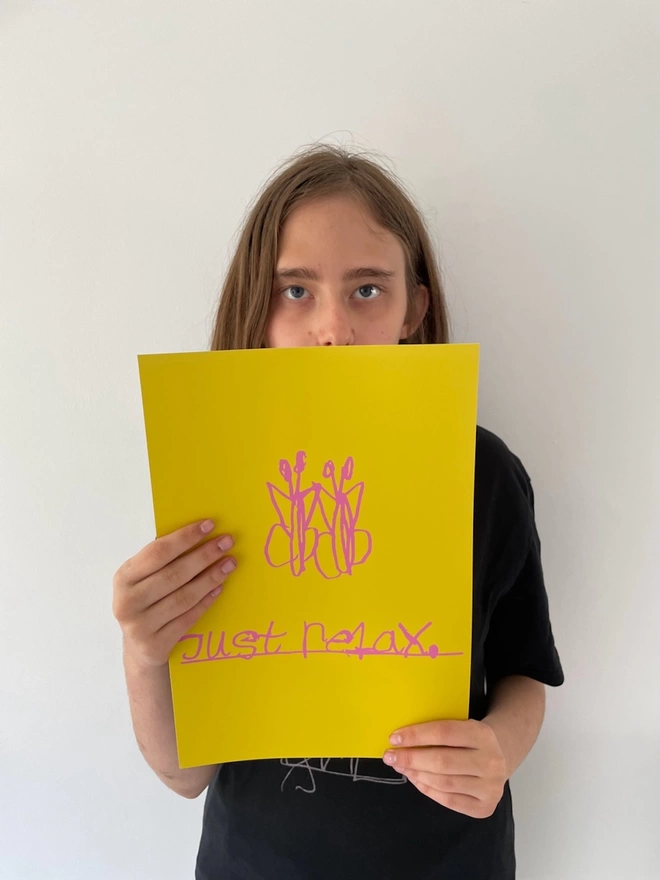 Piper is holding her digital print which is two pink butterflies and underneath them in her unique handwritten font are the works just relax underlined, on a yellow background.