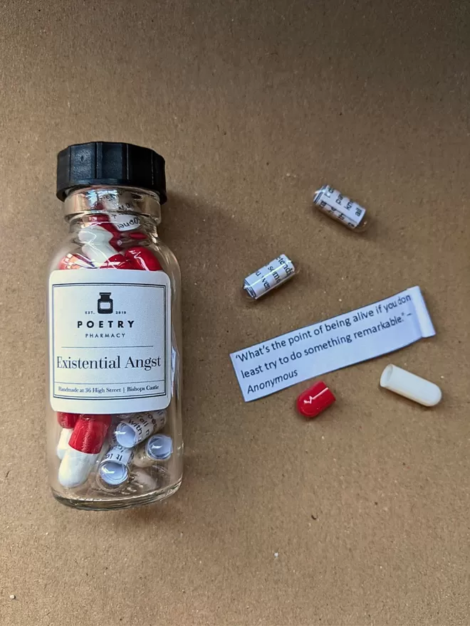 A glass bottle of Existential Angst poetry pills on a brown paper background, next to some loose capsules and a partially rolled up slip of paper with a poetry quote printed on it