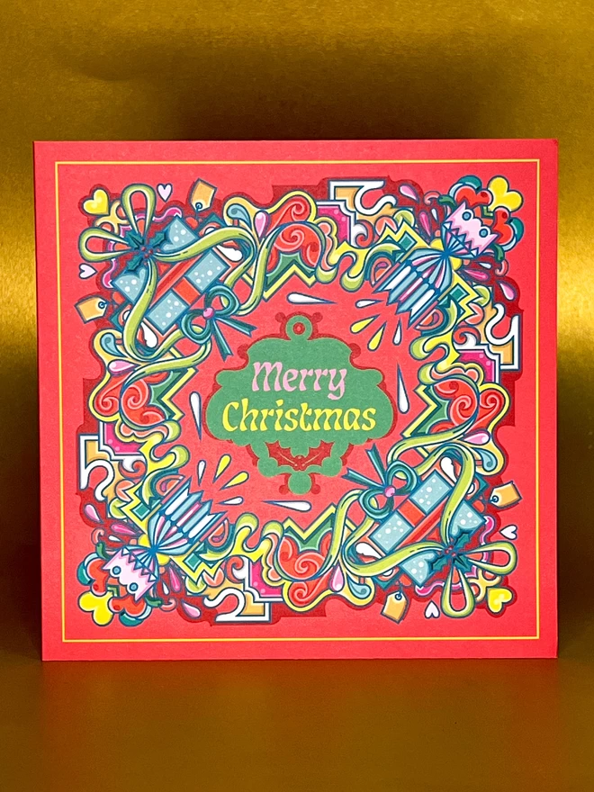 An abstract, square Christmas card design, with Merry Christmas in the centre, surrounded by a multi-coloured design including crackers and presents, on a red background, in front of a gold backdrop.