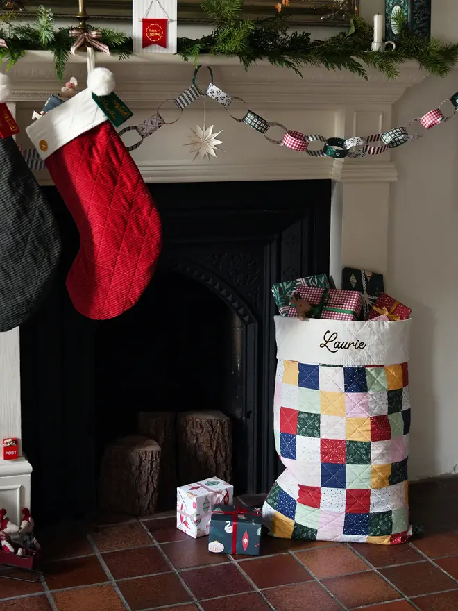 A handmade patchwork Christmas sack sits beside a black fireplace.