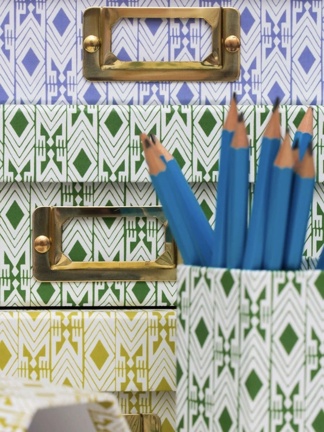 Tijan Hexagonal Brush Pot, a colourful patterned pencil pot sitting on a white surface accompanied by some folders. It is filled by some pencils. 