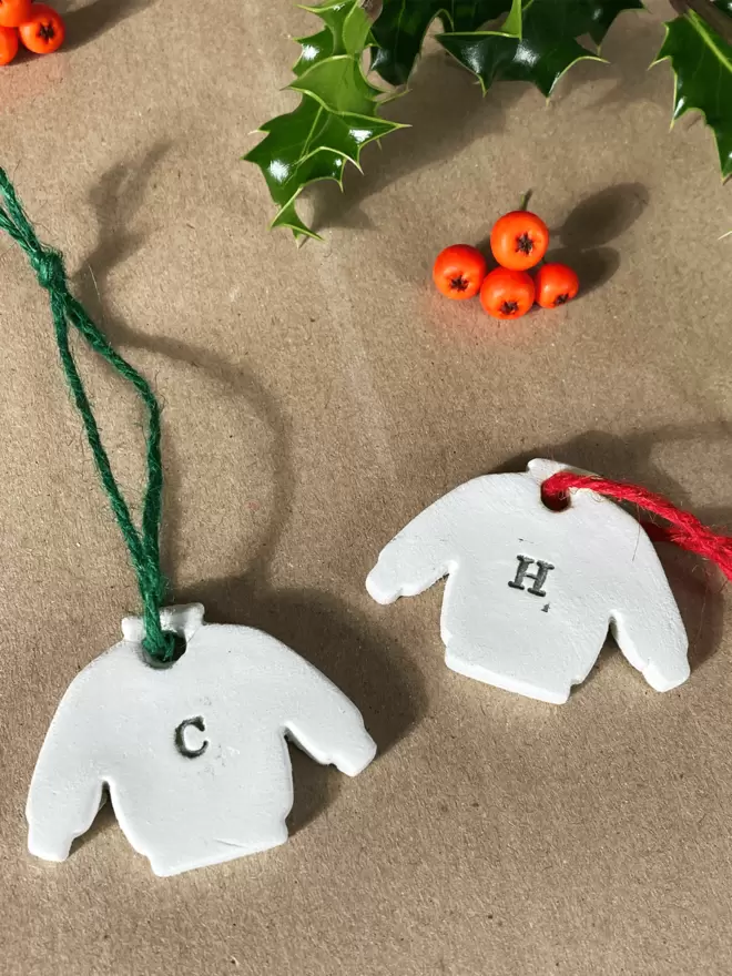 Two clay jumper tags, one with a 'C' and the other a 'H' in the centre of the jumper. One with green twine, the other with red twine. There is a sprig of holly in the background.