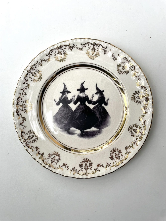 a coven of 3 witches dance hand in hand, the image is victorian in black and white and is printed directly onto an ornate vintage plate