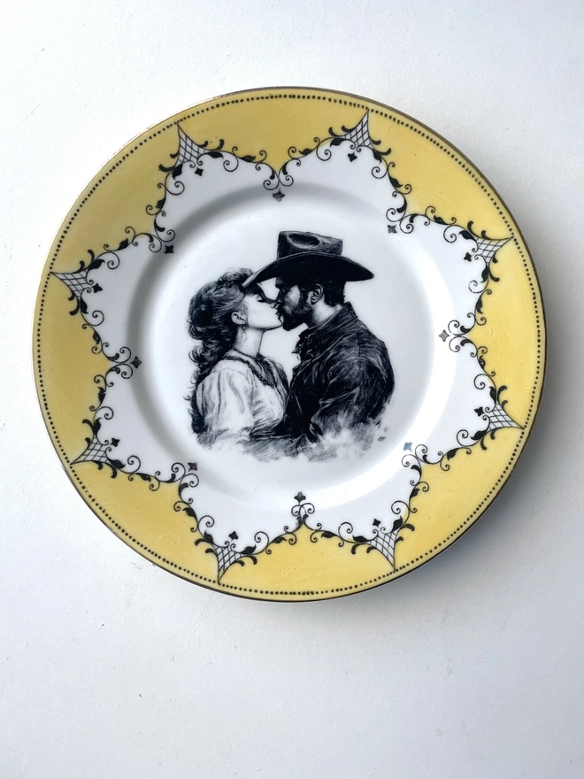 a vintage plate with an ornate border has a black and white vintage illustration of a cowboy and cowgirl sharing a kiss in the centre