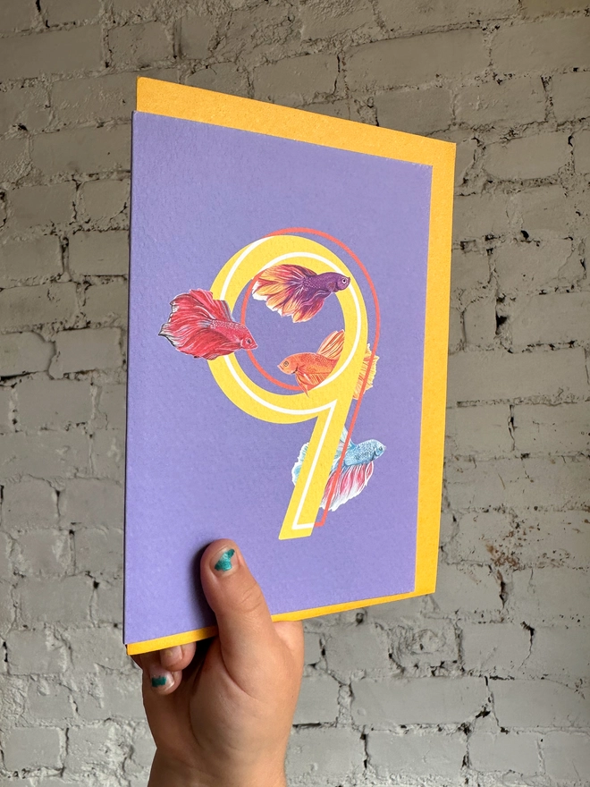 bright purple card with a big yellow number 9 and three bright illustrated betta fish with a bright yellow envelope