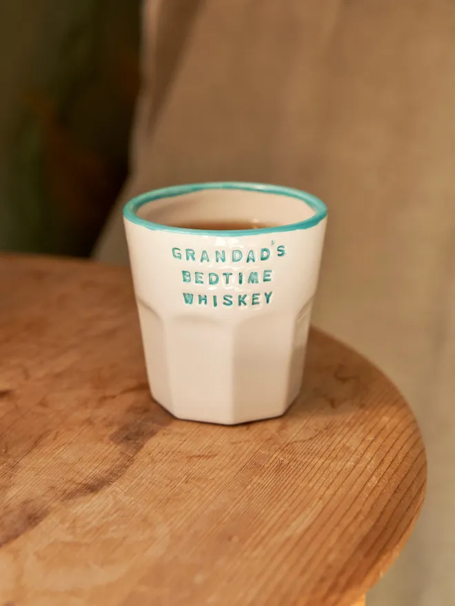 'Grandad's Bedtime Whiskey' in a teal font stamped on a ceramic cup with a teal rim, on a wooden table