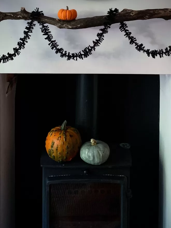 An orange and a green pumpkin on a black log burner below a black strinsel (string tinsel) decorated branch holding a smaller pumpkin against a white background