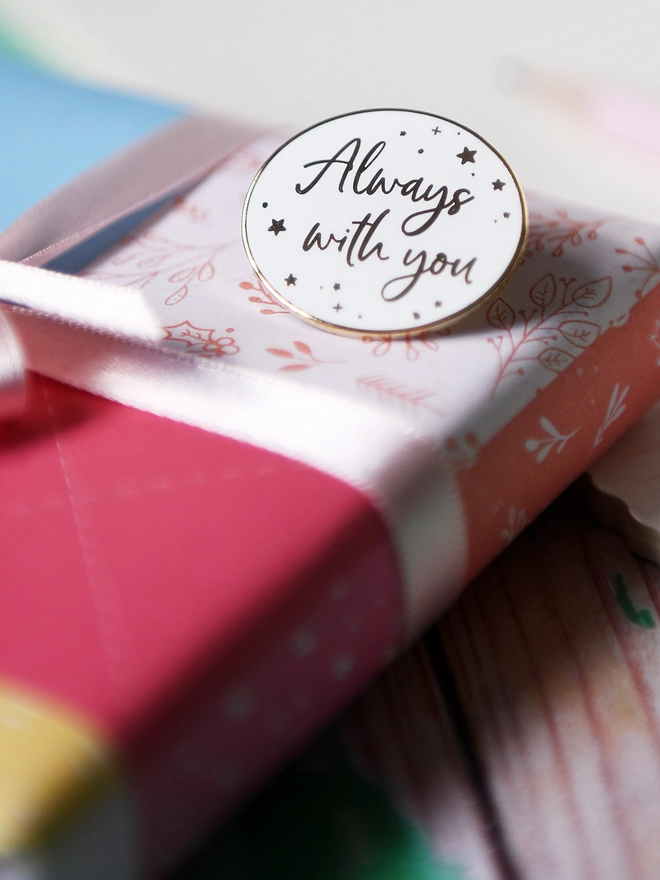 A white and gold enamel pin badge rests on a wrapped gift. It has a subtle star design and the words "Always with you".