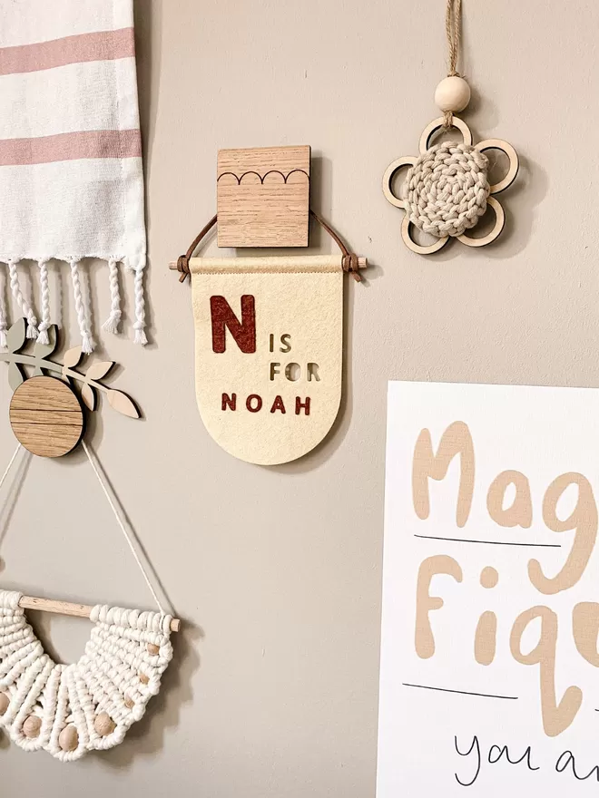 Yellow flag in the middle of a grey wall alongside other hanging objects. Small wooden flower, crochet semi circle all hung by small wooden hooks.