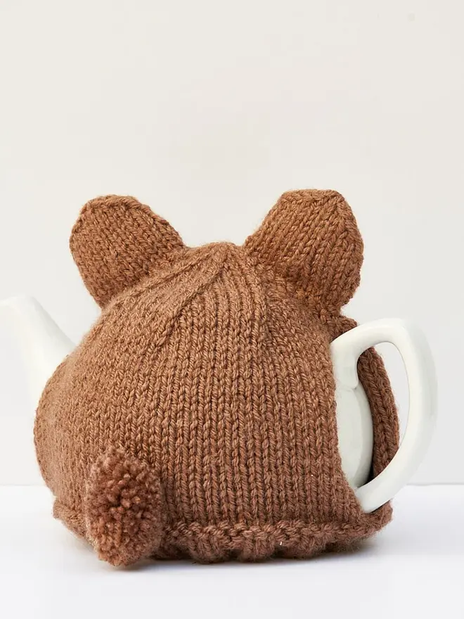 Brown Bear Tea Cosy Teddy Bears Picnic Treat, a brown bear tea pot cover sitting on a white tea pot, on a white surface against a grey backdrop. 