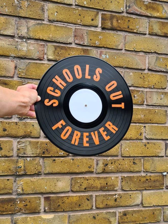 School's out forever in orange block text, on black vinyl record being held in front of brick wall