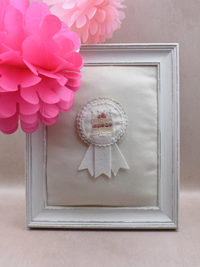 White and Beige felt rosette with embroidered Birthday Cake as the central design.  Pinned to a padded frame with paper decorations.