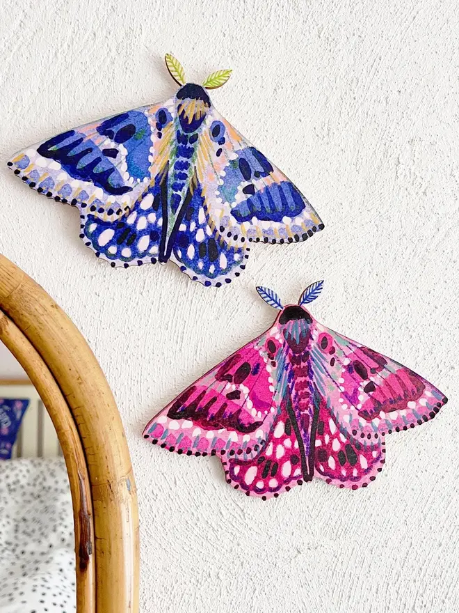 wooden moth wall decoration. pink and blue wooden moth decorations hung on plain white wall.