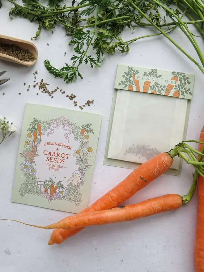A pale green "Carrot Seeds for Excellent Eyesight" card, framed by an illustrated bunny, chick, daffodils and carrots. Beside it is a glassine "Plant Me" seed pouch, with fresh carrots and their leafy tops.