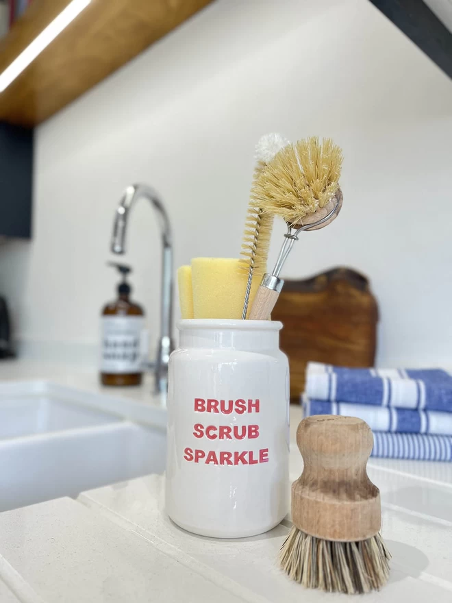 A handmade ceramic ‘Brush Scrub Sparkle’ Pot is holding an array of kitchen scrubbing brushes.