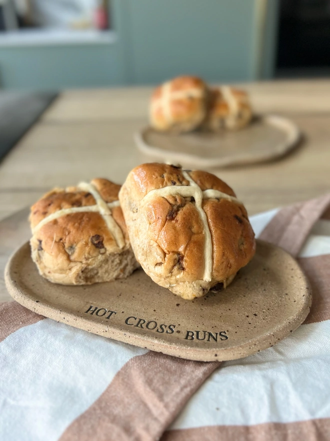 A oval plate with the wording 'hot cross buns' has two hot cross buns on it. The plate is in a specked brown clay