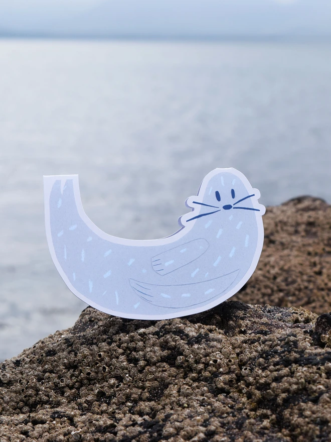 A cut out card of a seal, sitting on top of a rock with the sea in the background.