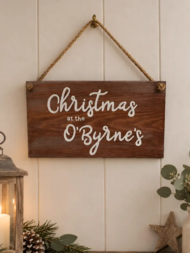 wooden personalised christmas sign hanging amongst christmas decorations and a candle. The sign says Christmas at the O'Byrne's.