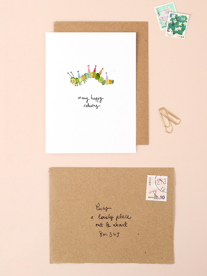 An image of a mini, rectangular white greetings card with an illustration of a cute colourful caterpillar wearing party hats across it's body with a hand written 'many happy returns' message seen under the central illustration. The mini greetings card is placed on top of a rectangular brown kraft envelope.