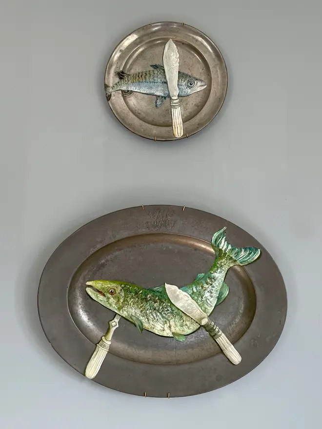 plate-scape. Oil Painted Fish Knife & Mackerel On Antique Pewter Plate, hanging on a grey wall, next to another plate. 