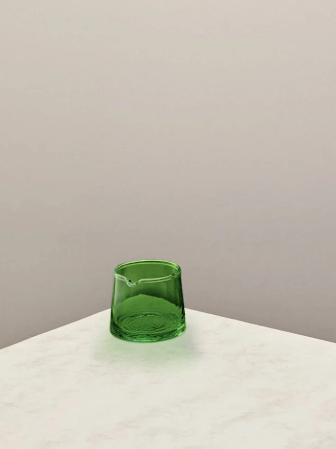 Low Jug, a small green glass jug sitting on a counter top against a plain backdrop. 