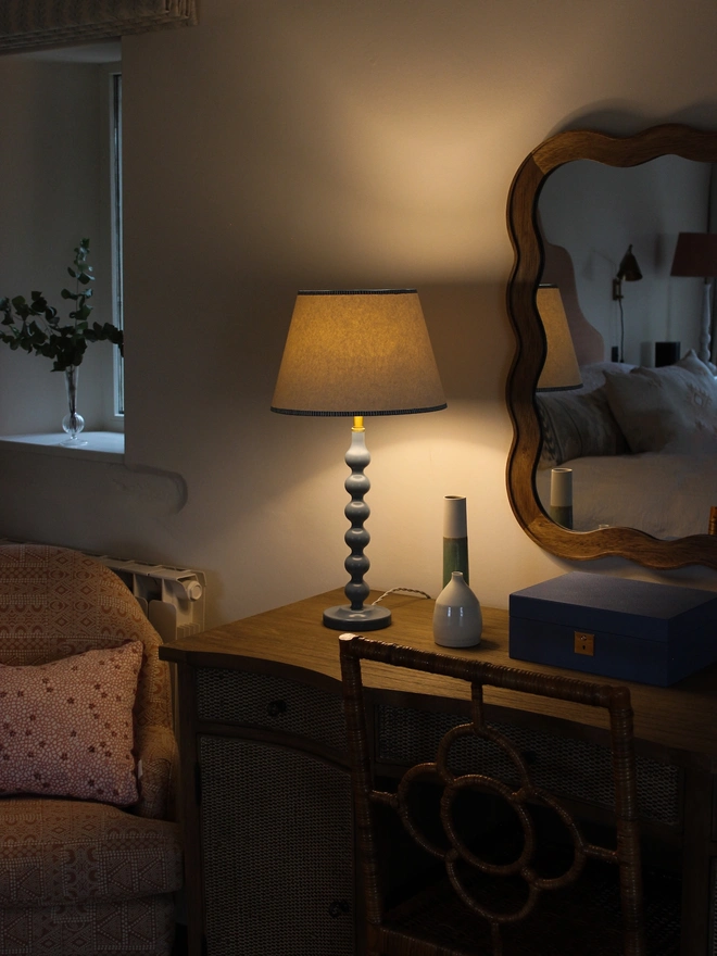 Blue table lamp with a white empire lampshade and blue striped trim, glowing warmly on a wooden dressing table beside a wavy wooden mirror and rattan chair in a cosy bedroom.