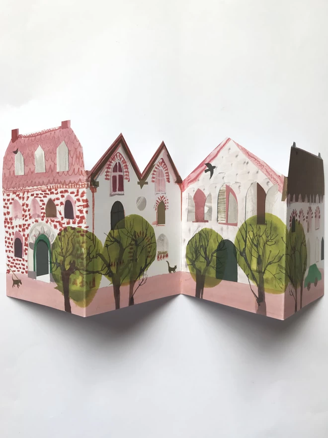 Folded concertina greetings card showing oink houses and green trees, resting on white background