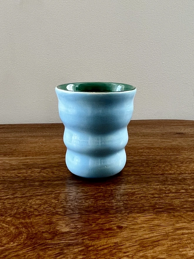 ceramic wavy tumbler with light blue glaze on the outside and dark green on the inside. the tumbler holds 250ml of liquid and sits on a wooden table