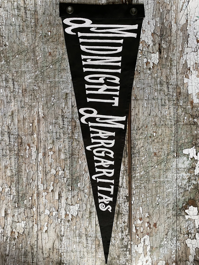A Black pennant flag hanging on a wall vertically with the words Midnight Margaritas in white.