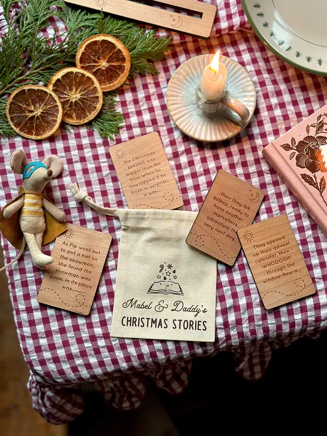 A personalised cotton story bag reading “Mabel & Daddy’s Christmas Stories” surrounded by engraved wooden story cards, dried oranges, greenery and a candle on a red gingham Christmas table.