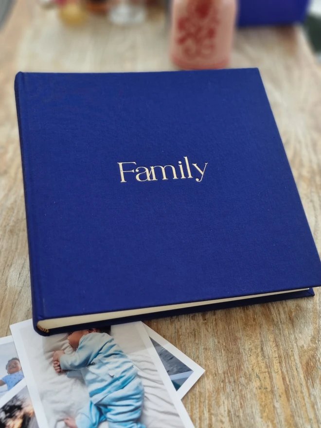 An indigo blue hand-bound photo album printed with the word 'Family' in gold lettering rests on a floor next to family photographs.