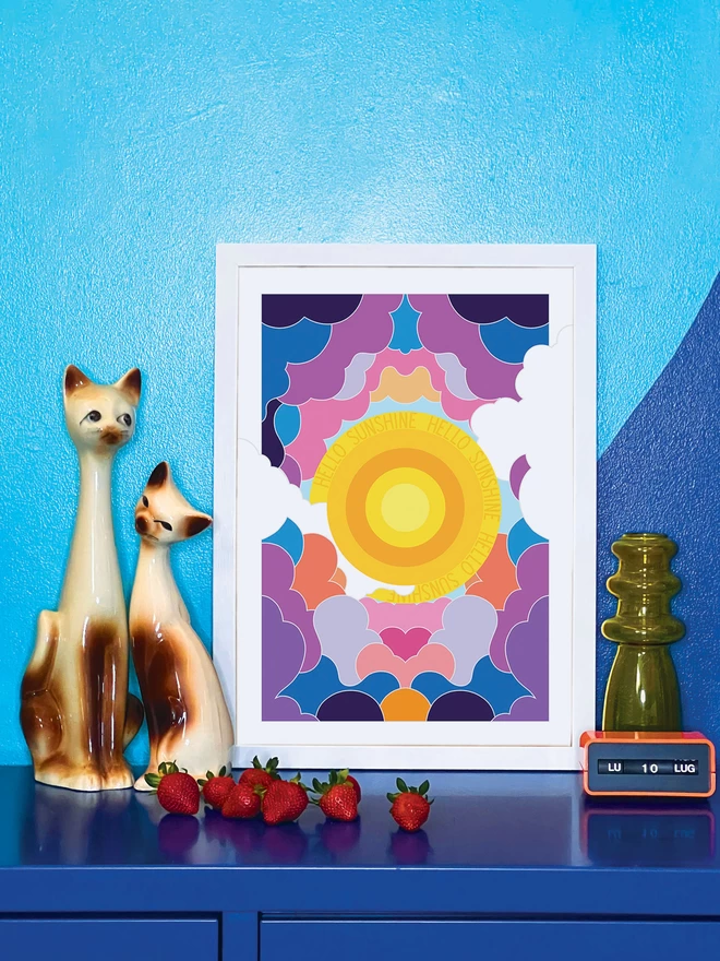 A concentric yellow circle sun with Hello Sunshine repeated three times in the outer ring sits at the centre of this portrait illustration. Surrounding it is an abstract design of clouds in purples, greys and oranges. The picture is in a white frame, against a turquoise and blue wall resting on a blue cabinet. Next to the picture are two cat ornaments, some ripe strawberries, a yellow glass vase and an orange Italian plastic calendar showing the date as ‘LU 10 LUG’.