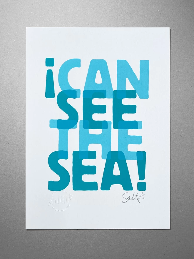 Full view of the A4 I Can See the Sea print. Two colours turquoise and teal, slightly overlapping words evoking the sense of waves. The print sits on a greyboard background.