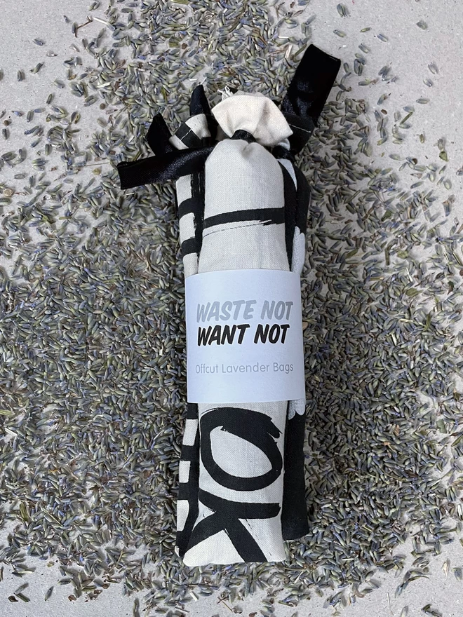 A bundle of three long style cotton lavender bags, tied at the tops, gathered with a branded cuff, sit on a scattered pile of fresh lavender - it says Waste Not Want Not, Offcut Lavender Bags. The fabric has bits of black and white printed pattern visible.