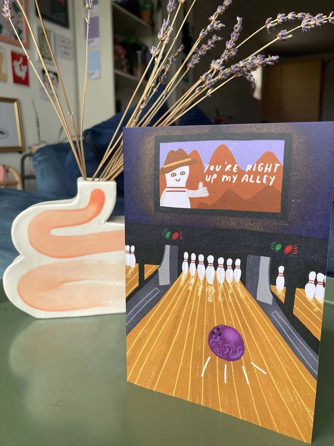 Illustrated greeting card on a side table with flowers behind. Card says 'you're right up my alley', and is an image of a bowling lane.
