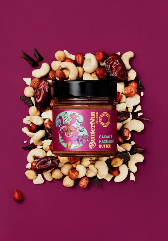 cacao and hazelnut butter. a jar of nut butter photographed against a magenta background, surrounded by assorted nuts and dried fruits.