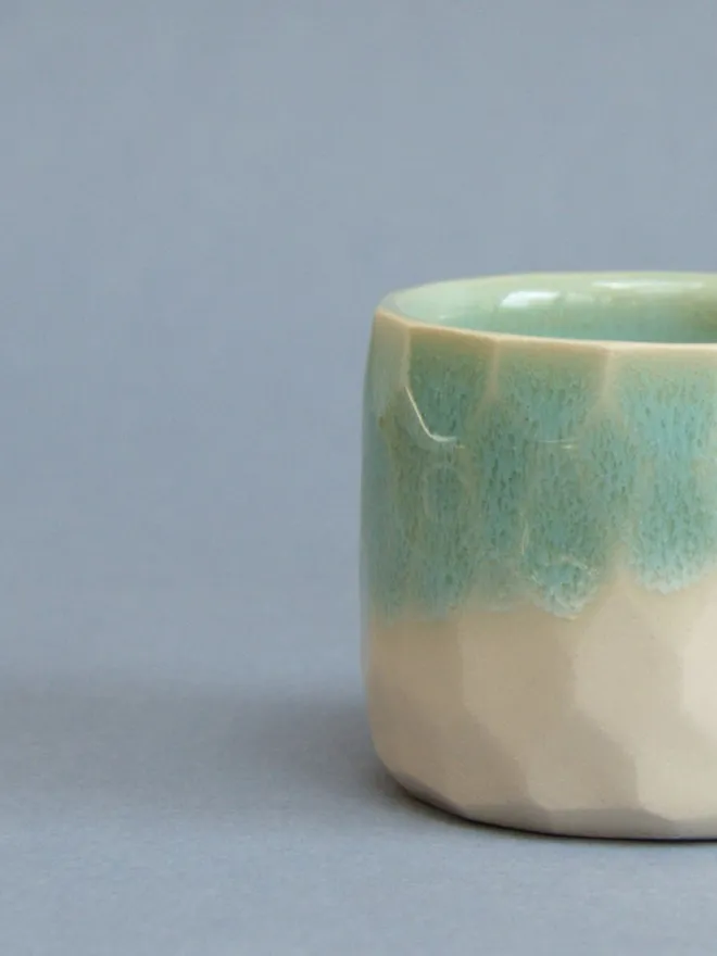  A close up of a straight sided cup which is half in view.  The cup has a turquoise top half and a glossy cream bottom half and the surface is decorated with a honeycomb hexagonal texture.