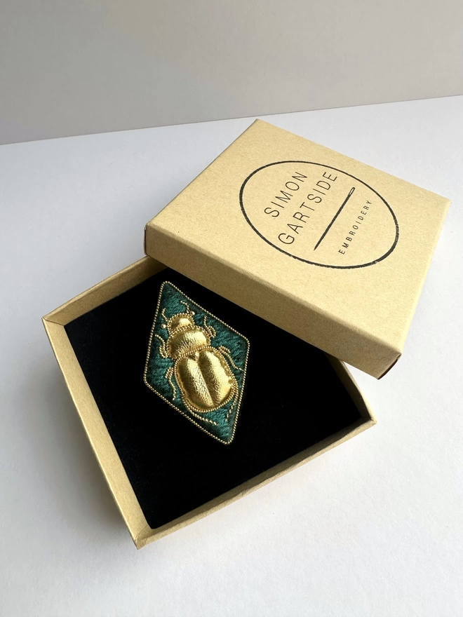 Diamond shaped brooch with golden beetle on a green background sat in an open box 