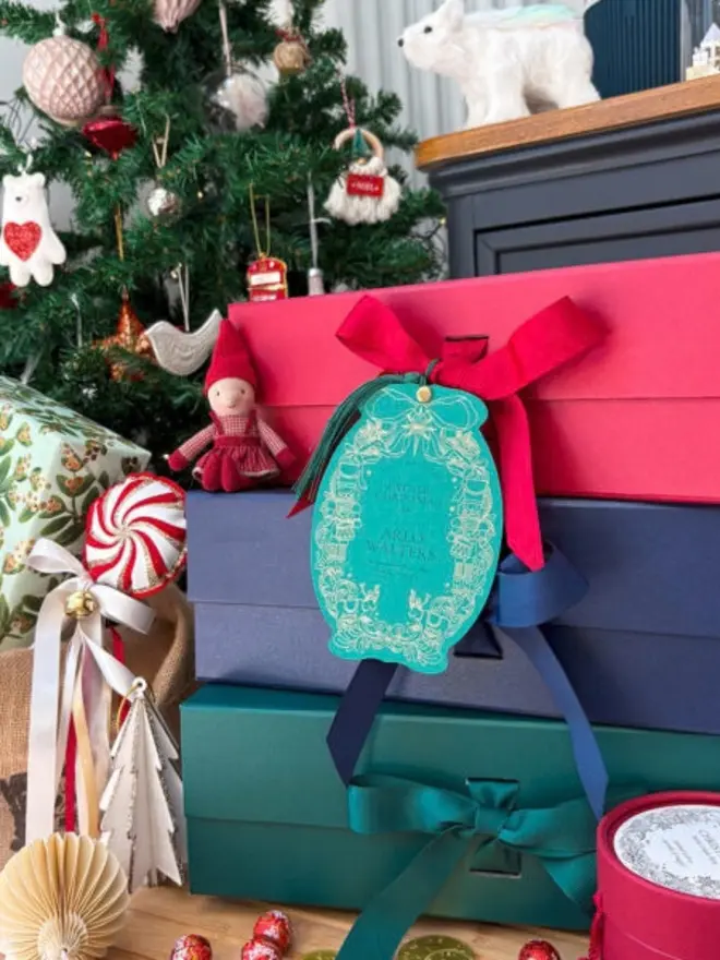 A personalised teal gift tag with a gold foil nutcracker and reindeer border and a matching tassel, hanging from a red ribbon on a stack of red, navy and green gift boxes. Around it are a decorated Christmas tree, an elf doll, a peppermint ornament and a polar bear figure.