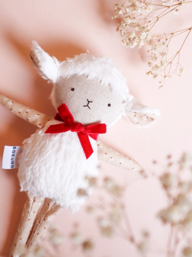 Handmade lamb doll with a fluffy white body, stitched face, beige floral fabric arms and legs, and a small red velvet bow tied around its neck.