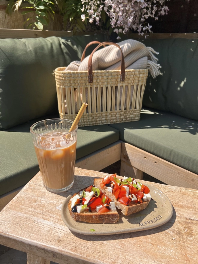 garden set up with a basket sat on a sofa with an iced coffee and an oval ceramic plate sat on a wooden table. Plate has the word 'Alfresco' stamped around the corner. 