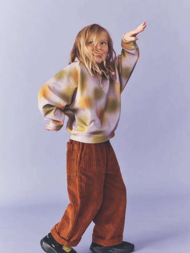 Child stands with hands raised against a lilac backdrop, wearing a Studio Koter lilac and ochre blurred-print collared sweatshirt with rust corduroy trousers.