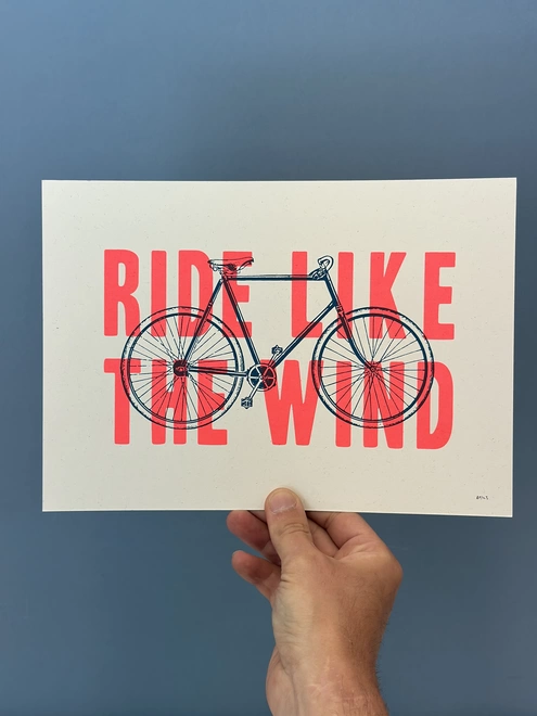 Photo of a hand holding an A4 Screen print showing an illustration of a traditional shaped bicycle with the words ride like the wind over the top in flouro pink typography