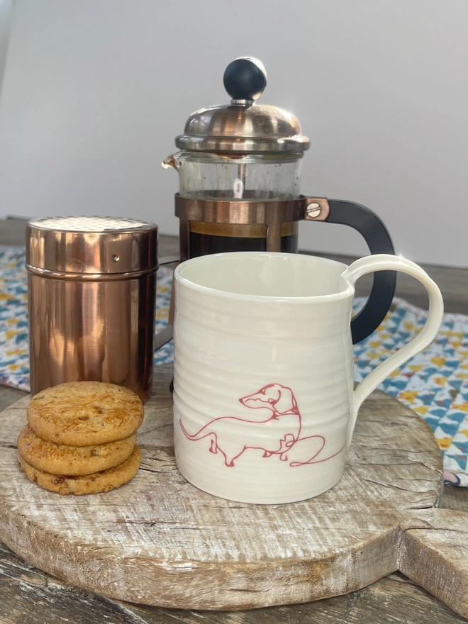 hand thrown porcelain mug with a red dachshund line drawing design on the side. Mug sits on table top amidst coffee scene with coffee pot and biscuits