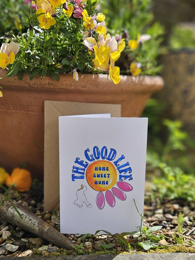 A white card with an illustration of a pink flower and a white bird. The Good Life is written using a retro font style in blue. In the centre of the flower it says Home Sweet Home. 